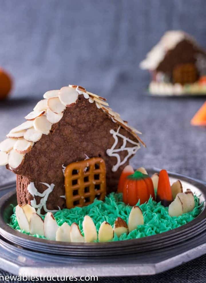 House-shaped chocolate brownie topped with shaved almond roof shingles and fence. The house is deocrated with candy corn, a pretzel door and green icing grass.