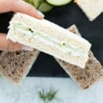 hand holding cucumber sandwich above cucumber slices and more sandwiches