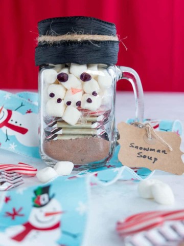 Mason jar decorated as a snowman with hot chocolate mix, candy cane, marshmallows and Ghirardelli peppermint bark squares inside.