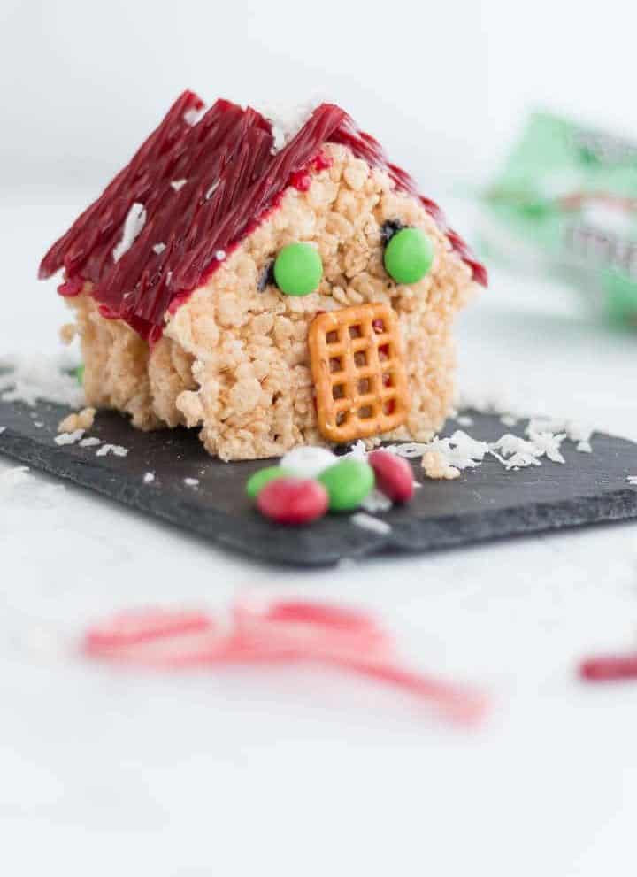 edible 3D house made of christmas rice krispie treats, red licorice roof shingles, mint M&M windows, pretzel doorway, and coconut snowflakes.